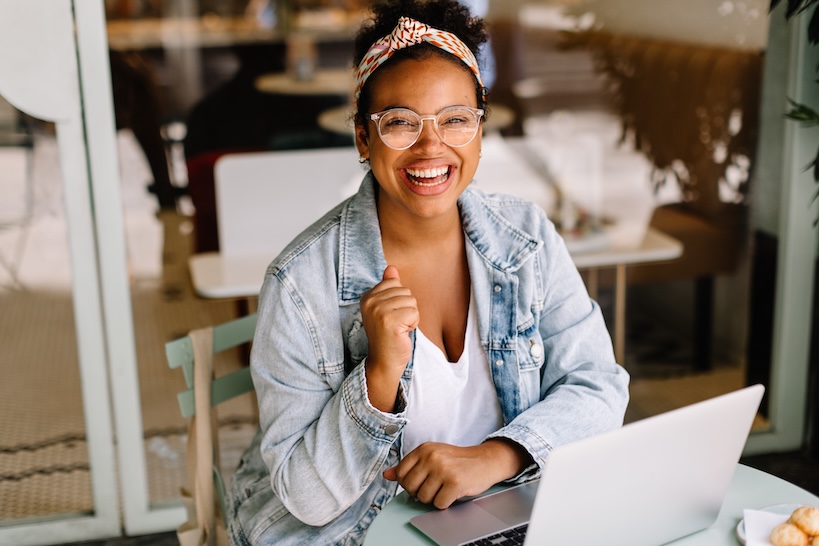 A woman wearing a denim jacket, clear glasses, and a colorful headband, with a
big smile sitting behind her laptop