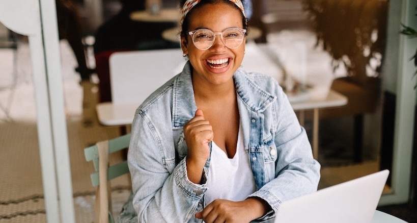 A woman wearing a denim jacket, clear glasses, and a colorful headband, with a big smile sitting behind her laptop