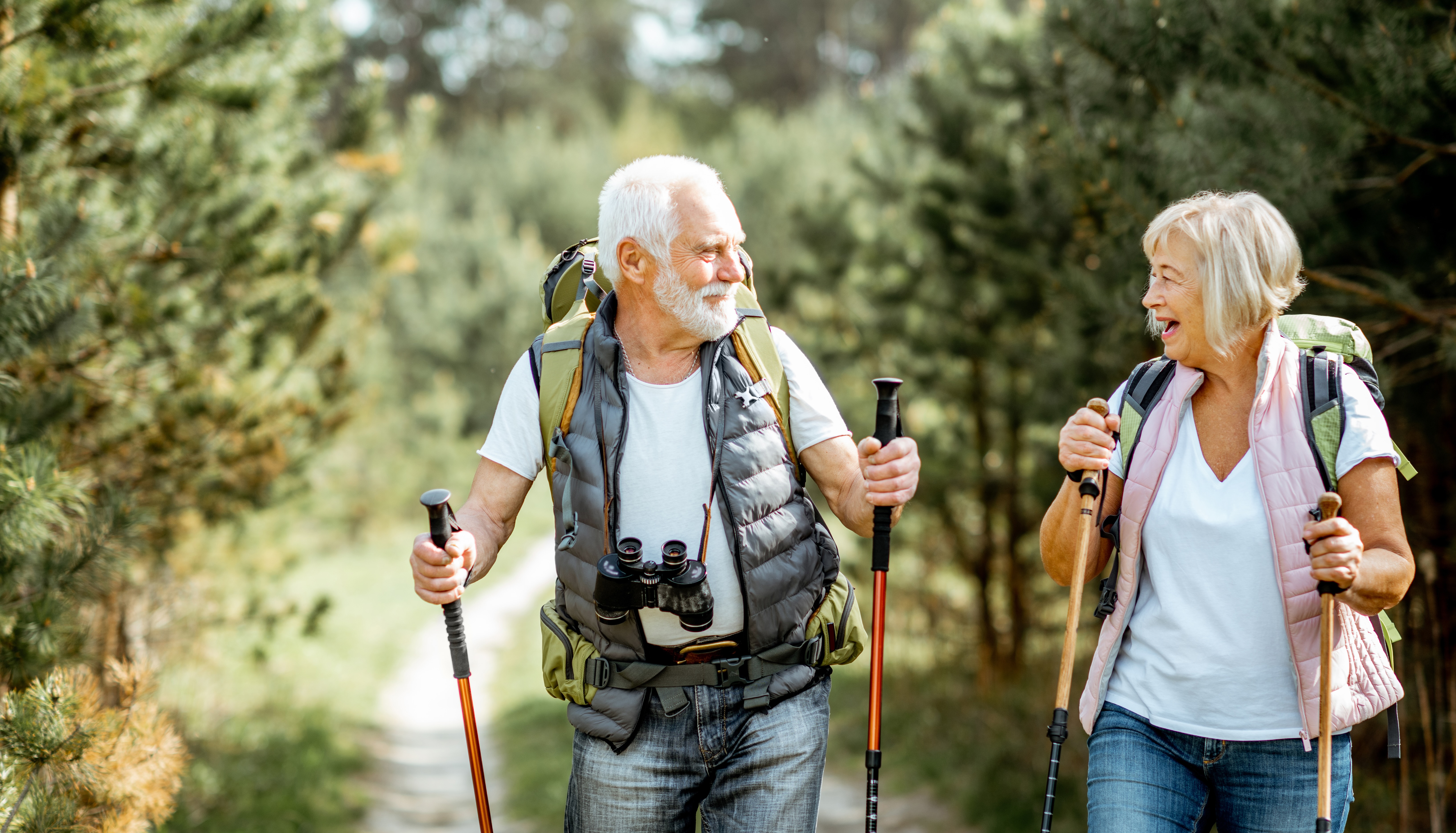 Happy older couple smiling while hiking with their trekking sticks and backpacks through a forest
