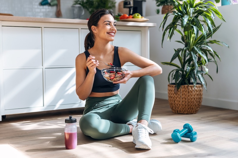 Athletic woman eating a healthy bowl of fruit sitting on floor in the kitchen
at home.
